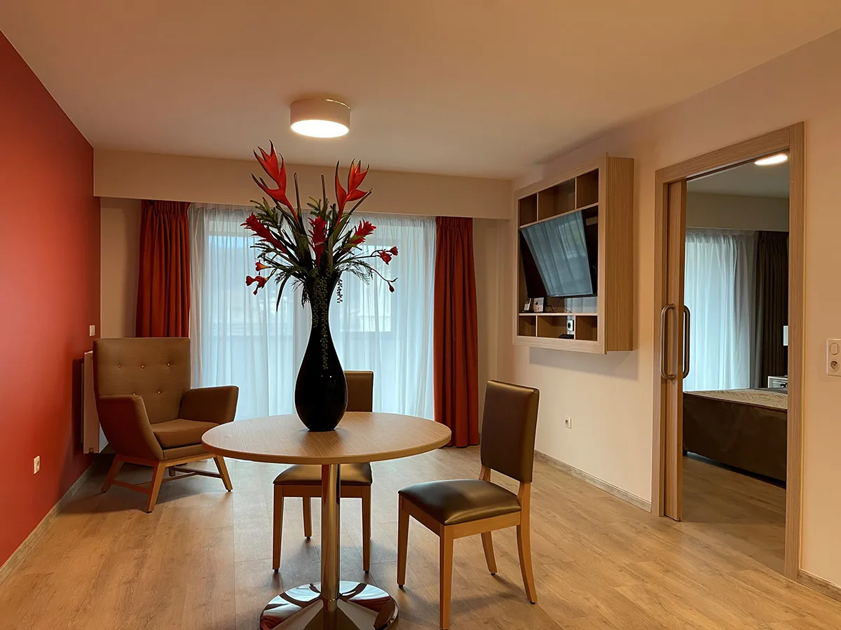 Séjour d'un appartement de la résidence seniors OBEO d'Aix-les-Bains : mur accent rouge, table ronde fleurie, fauteuil et chambre visible en enfilade