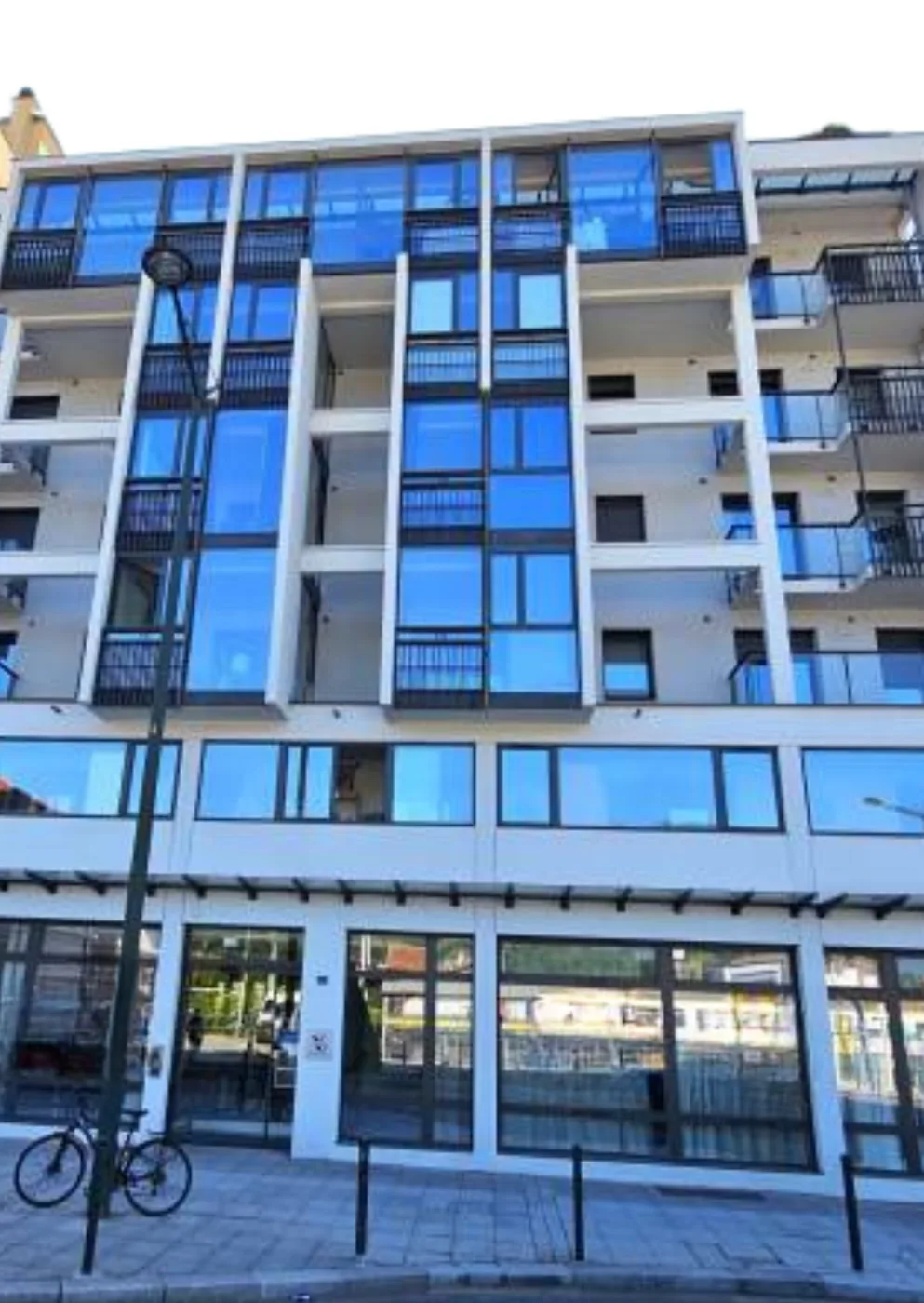 Façade de la résidence OBEO d’Aix-les-Bains vue de la rue : six étages de balcons vitrés bleutés, commerces en rez-de-chaussée et vélo garé le long du trottoir