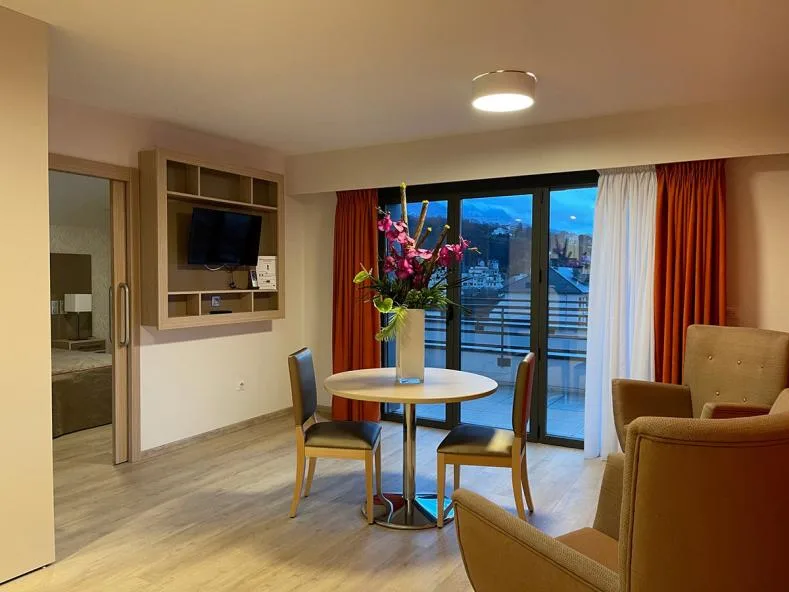 Séjour d’un appartement témoin d’Aix-les-Bains : table ronde fleurie, fauteuils, téléviseur encastré et baie vitrée ouvrant sur le balcon et les montagnes