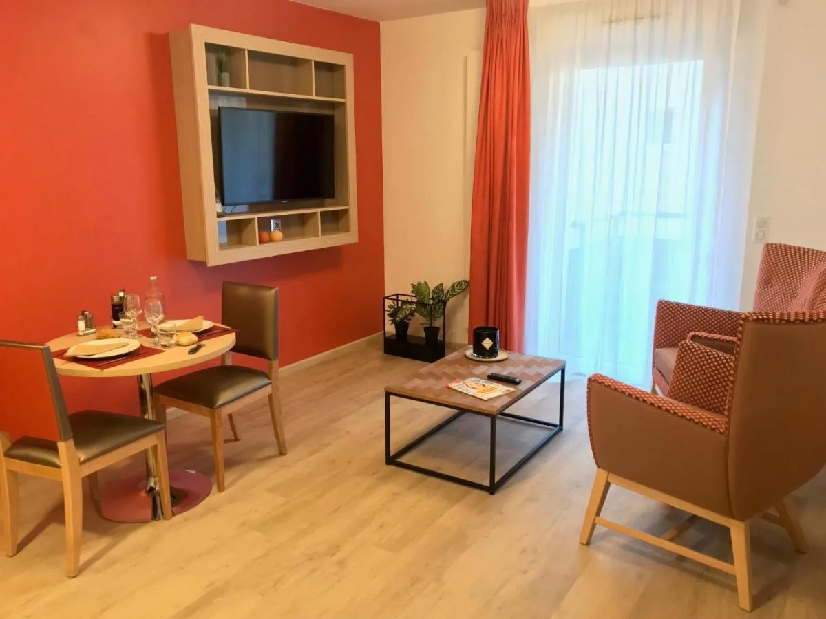 Living area of a show apartment in Chambéry: red accent wall, television set in a wooden alcove, laid dining table and armchairs by the window