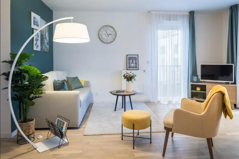 Séjour meublé d’un appartement OBEO : canapé clair, fauteuil jaune, plantes et lampe arceau