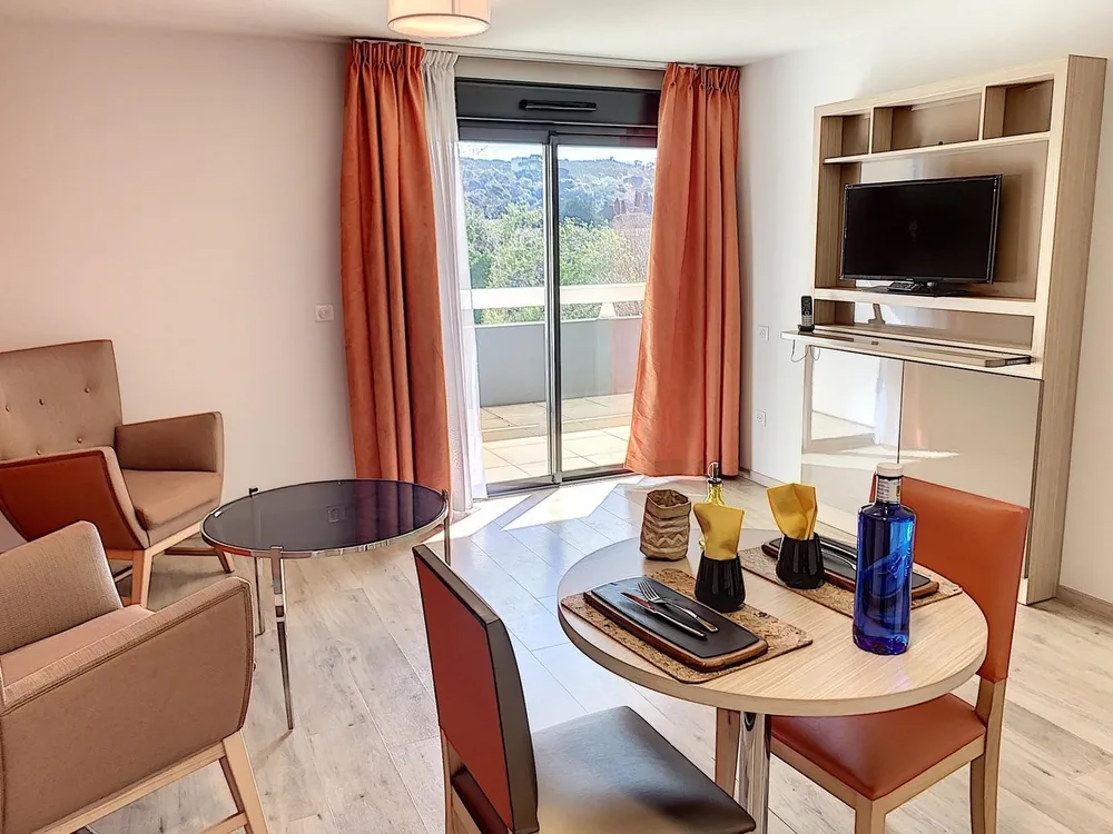 Séjour d’un appartement témoin du Cannet : fauteuils, table ronde dressée, téléviseur et baie vitrée ouvrant sur le balcon et les collines