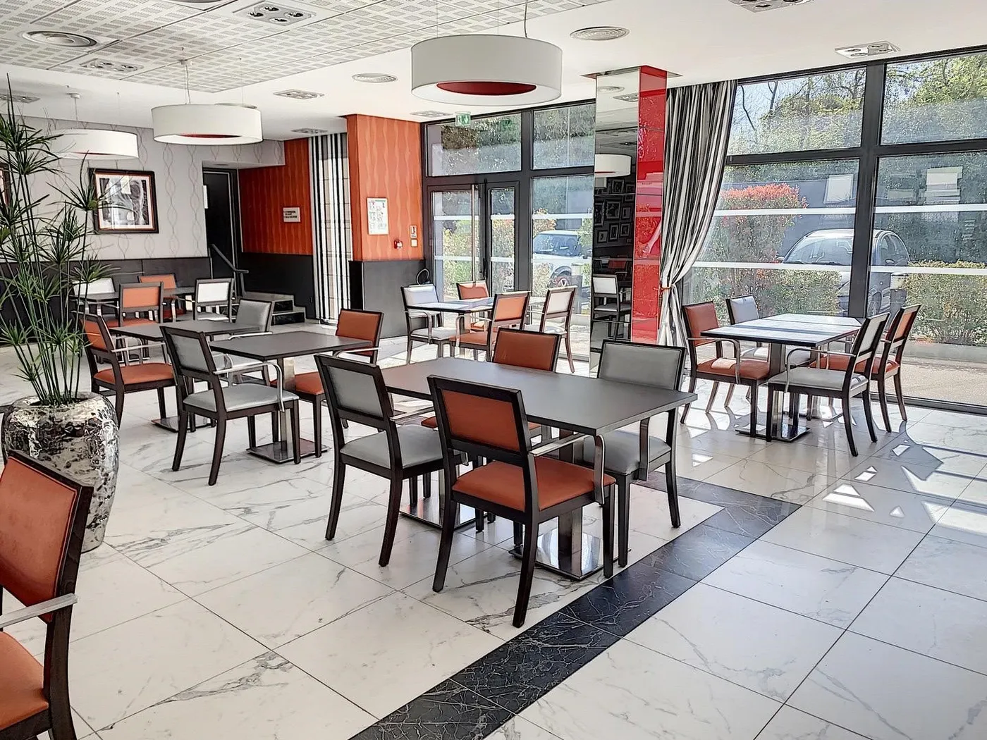 Restaurant de la résidence seniors OBEO du Cannet : salle lumineuse aux baies vitrées toute hauteur, tables dressées et chaises orange sur sol en marbre