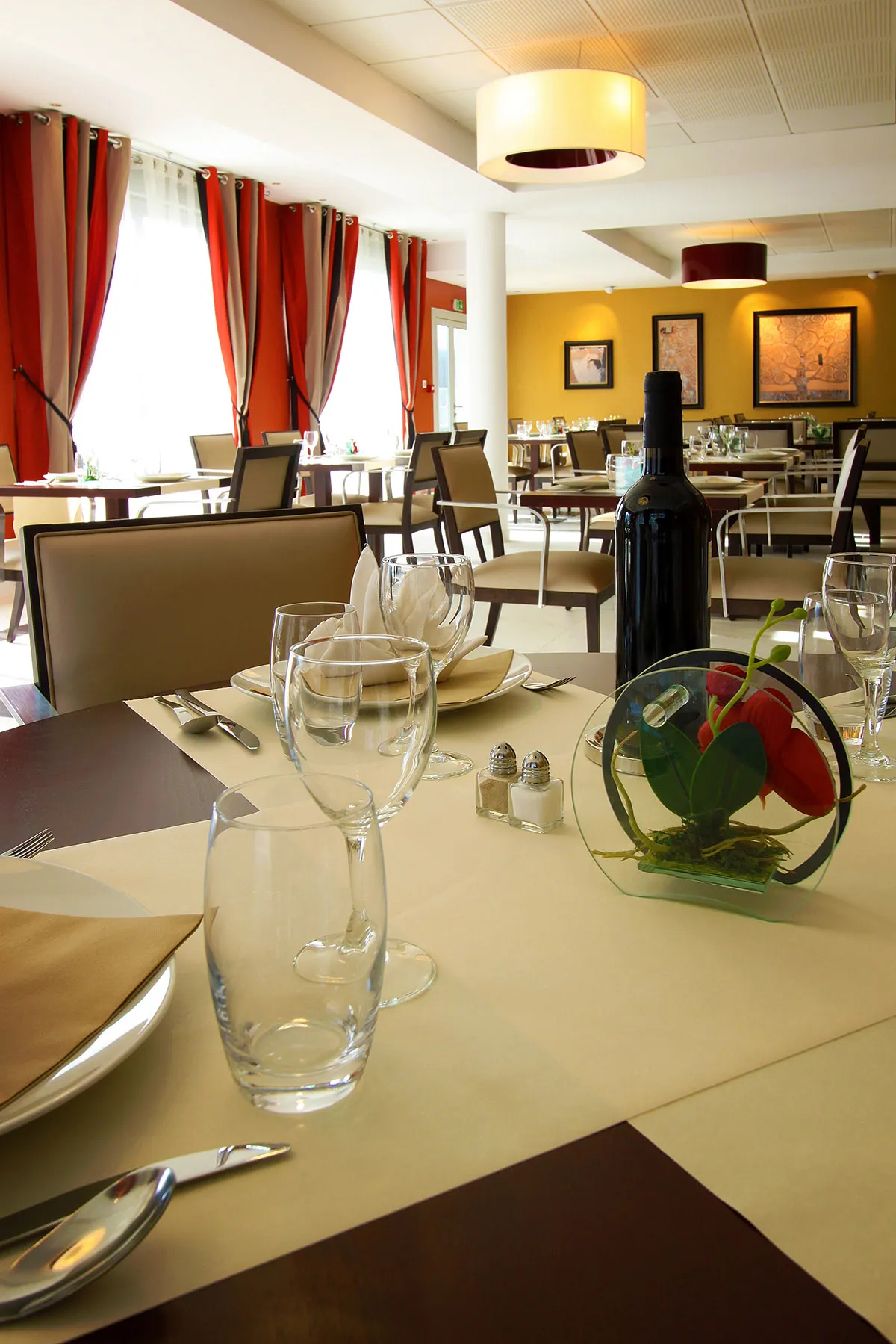 Salle de restaurant de la résidence seniors OBEO de Montreuil : tables dressées avec verres à pied, baies vitrées aux rideaux rayés rouges et murs jaunes ornés de tableaux