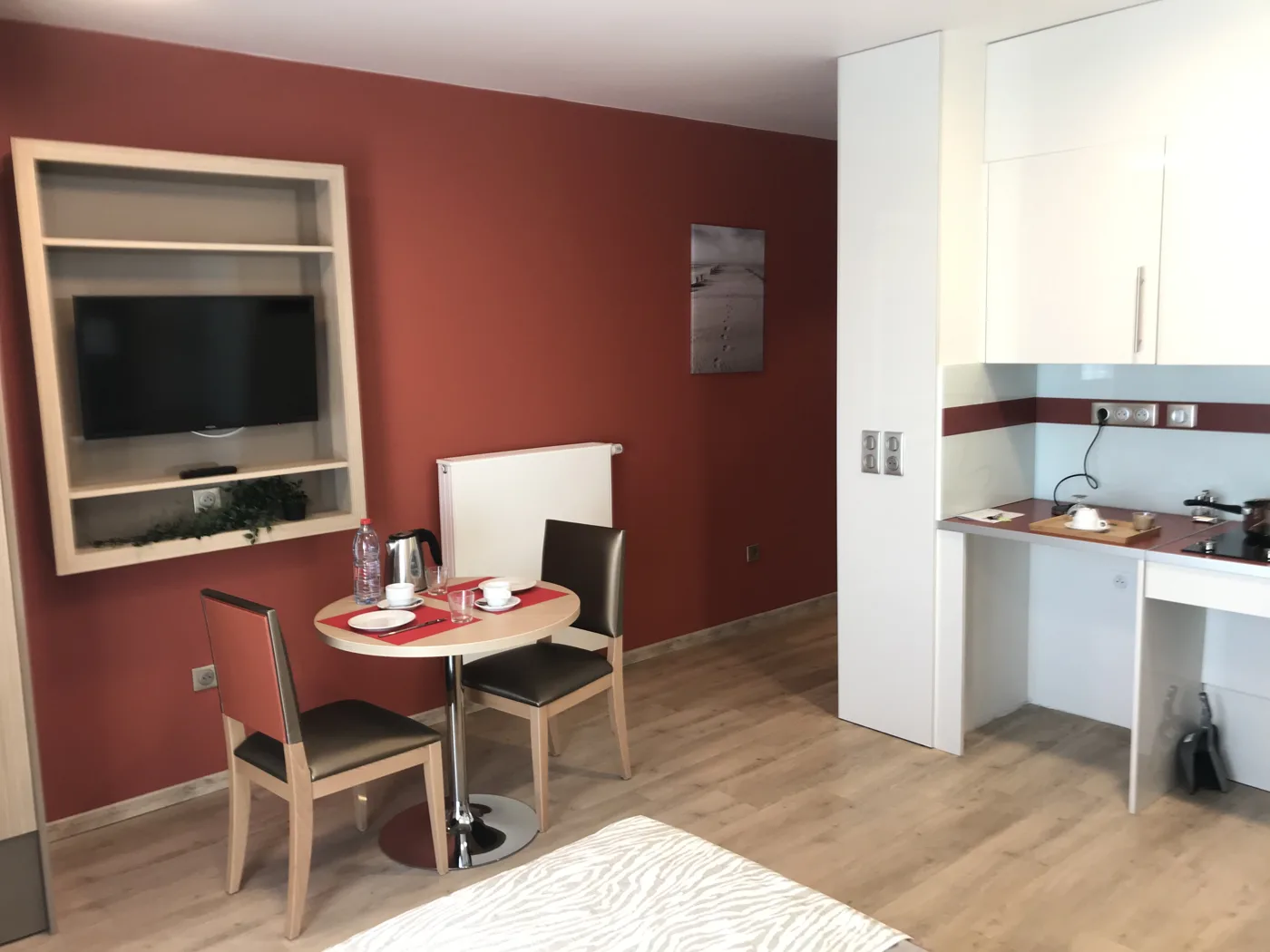 Appartement témoin de Montreuil : mur accent terracotta, téléviseur encadré de bois, table de repas pour deux et kitchenette équipée