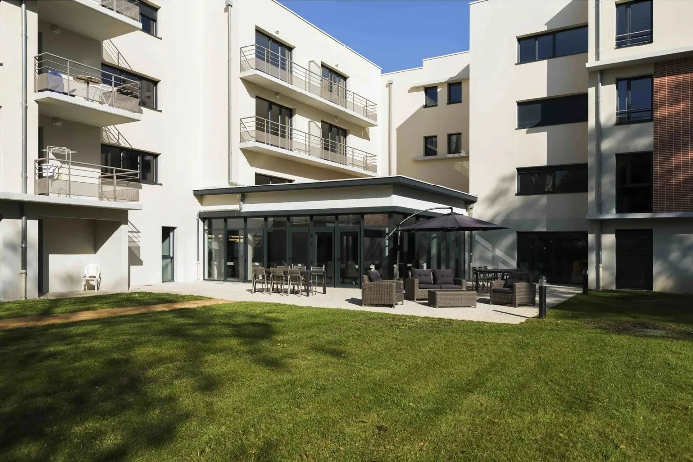 Jardin ensoleillé de la résidence services seniors OBEO à Nevers : terrasse avec salon de jardin et parasol devant la véranda vitrée, au pied des appartements avec balcon