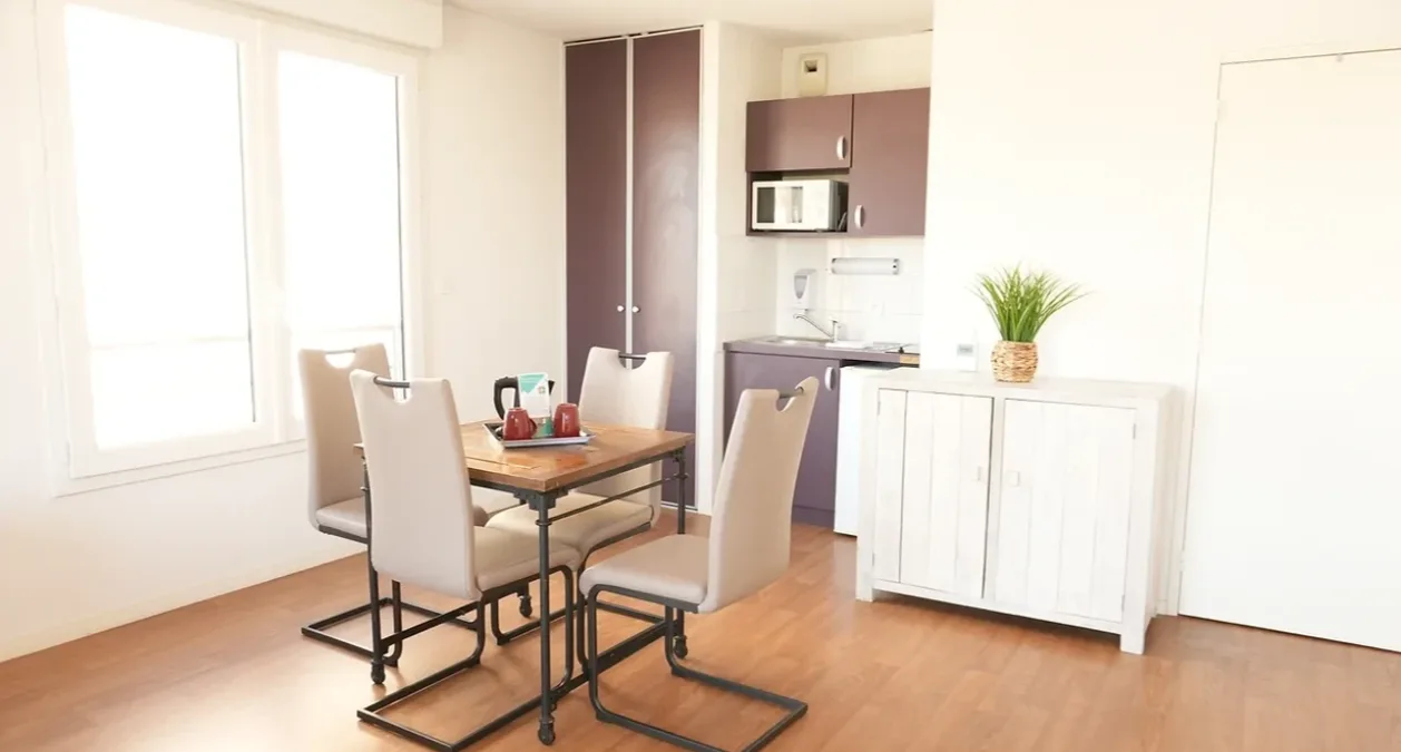 Coin repas d’un appartement témoin de Nevers : table en bois et chaises blanches devant la kitchenette et le buffet, sous une grande fenêtre