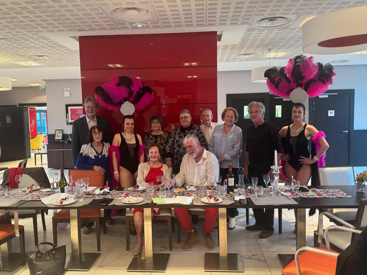 Photo de groupe autour de la table de fête : résidents, proches et équipe encadrés par les deux danseuses de cabaret