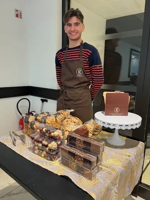Un jeune chocolatier en tablier rayé se tient derrière son étal de pâtisseries et de chocolats