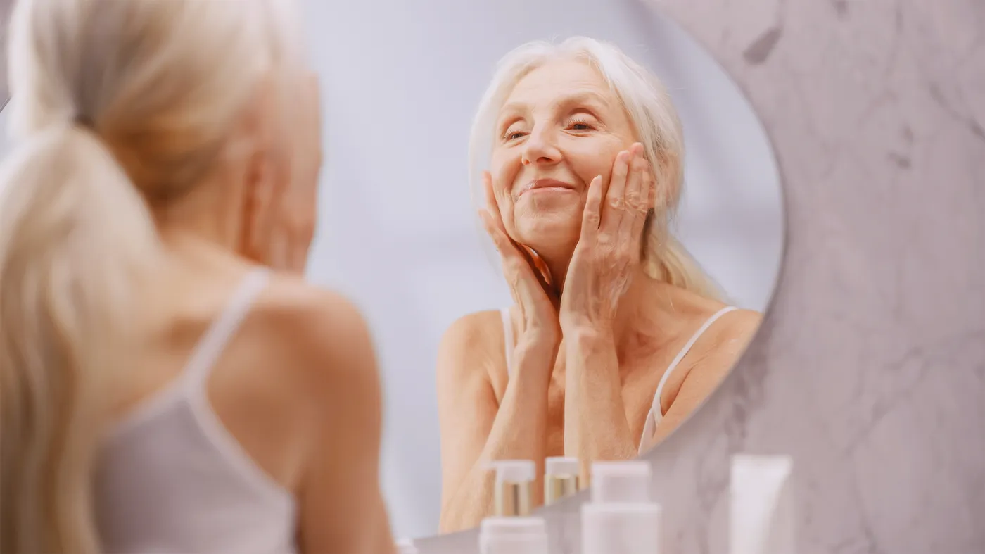 Une femme âgée se masse le visage du bout des doigts devant un miroir