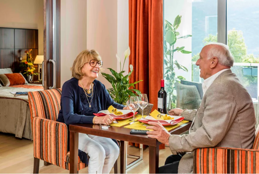 Un couple de seniors partage un repas à la table de son appartement, le lit et la baie vitrée derrière eux