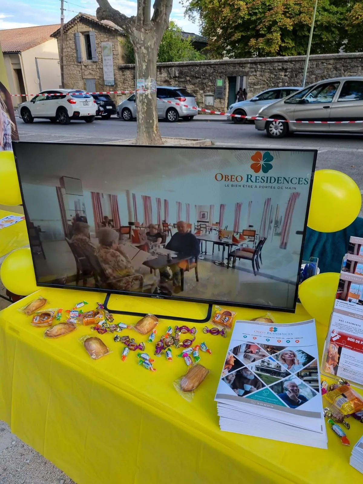 Le stand OBEO installé sous les arbres, avec son écran, ses brochures et sa nappe jaune