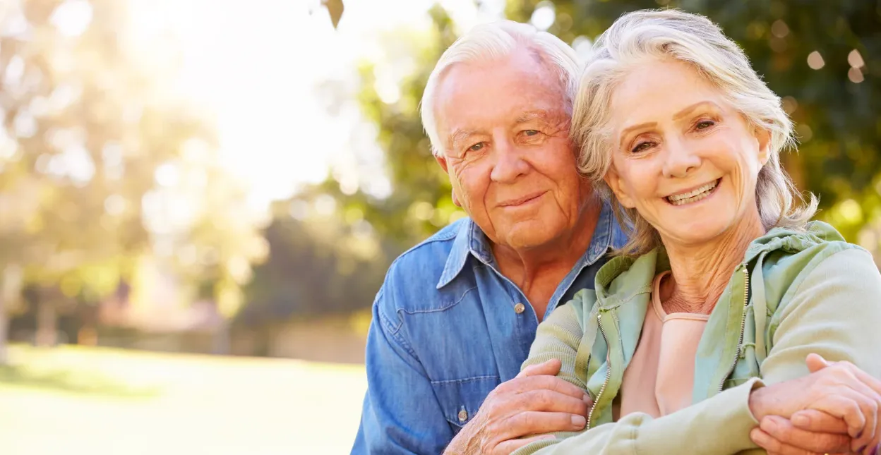 Un couple de seniors enlacé sourit face à l'objectif, des arbres ensoleillés en arrière-plan