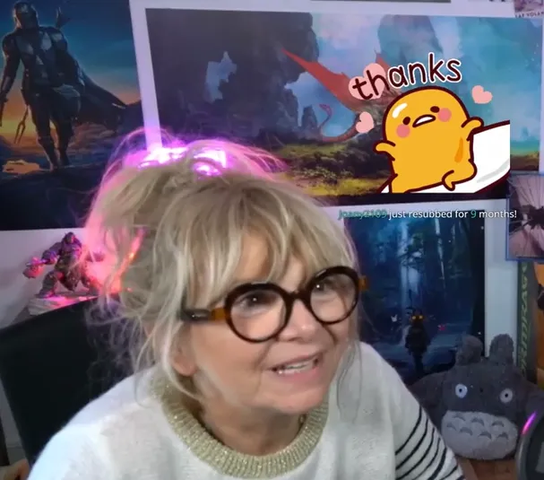 Une femme âgée à lunettes rouges diffuse une partie de jeu vidéo en direct, un message de remerciement affiché à l'écran derrière elle