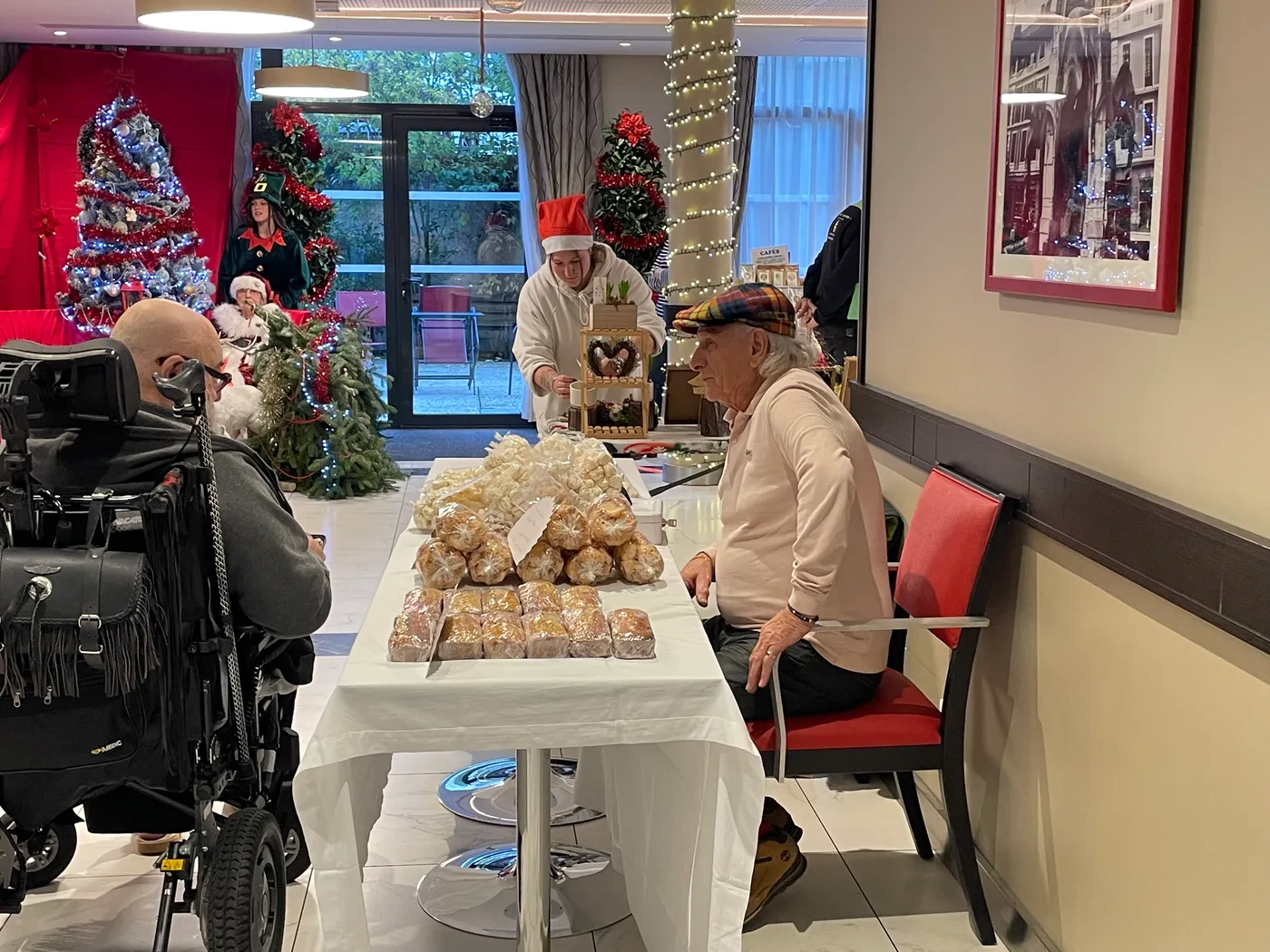 Un stand de brioches emballées empilées sur une table blanche, tenu par un membre de l'équipe coiffé d'un bonnet de Père Noël, devant des résidents