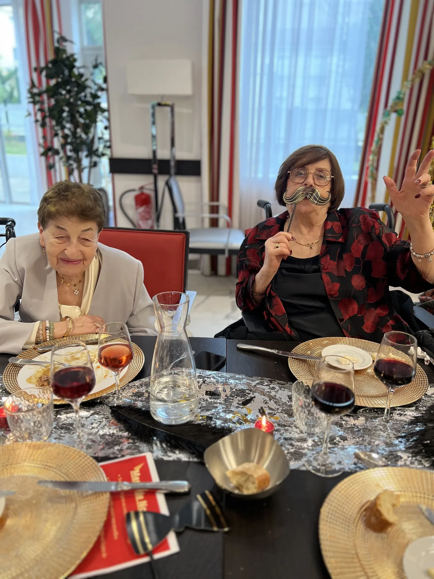Deux résidentes à table, l'une faisant un signe de la main, avec assiettes dorées et verres de vin
