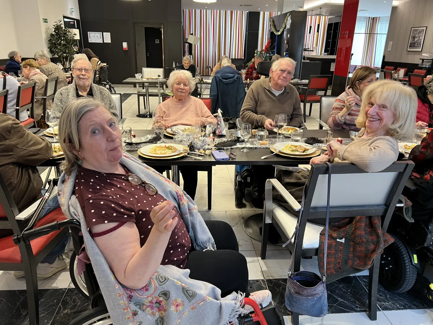 Des résidents installés aux tables rondes de la salle à manger, l'une d'elles souriant à l'objectif
