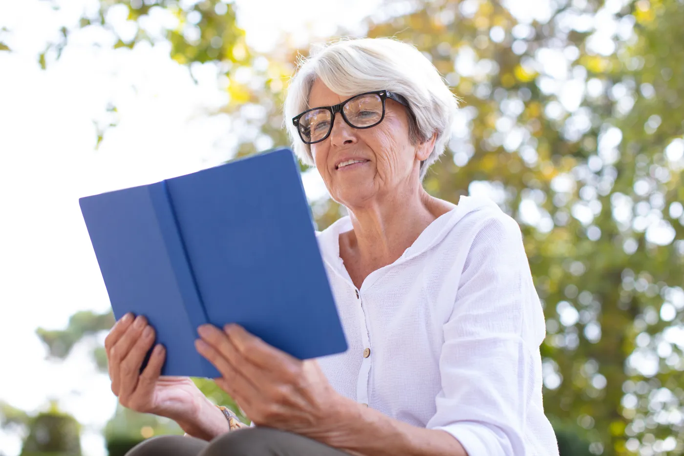 Une femme âgée assise dehors, un livre bleu ouvert entre les mains