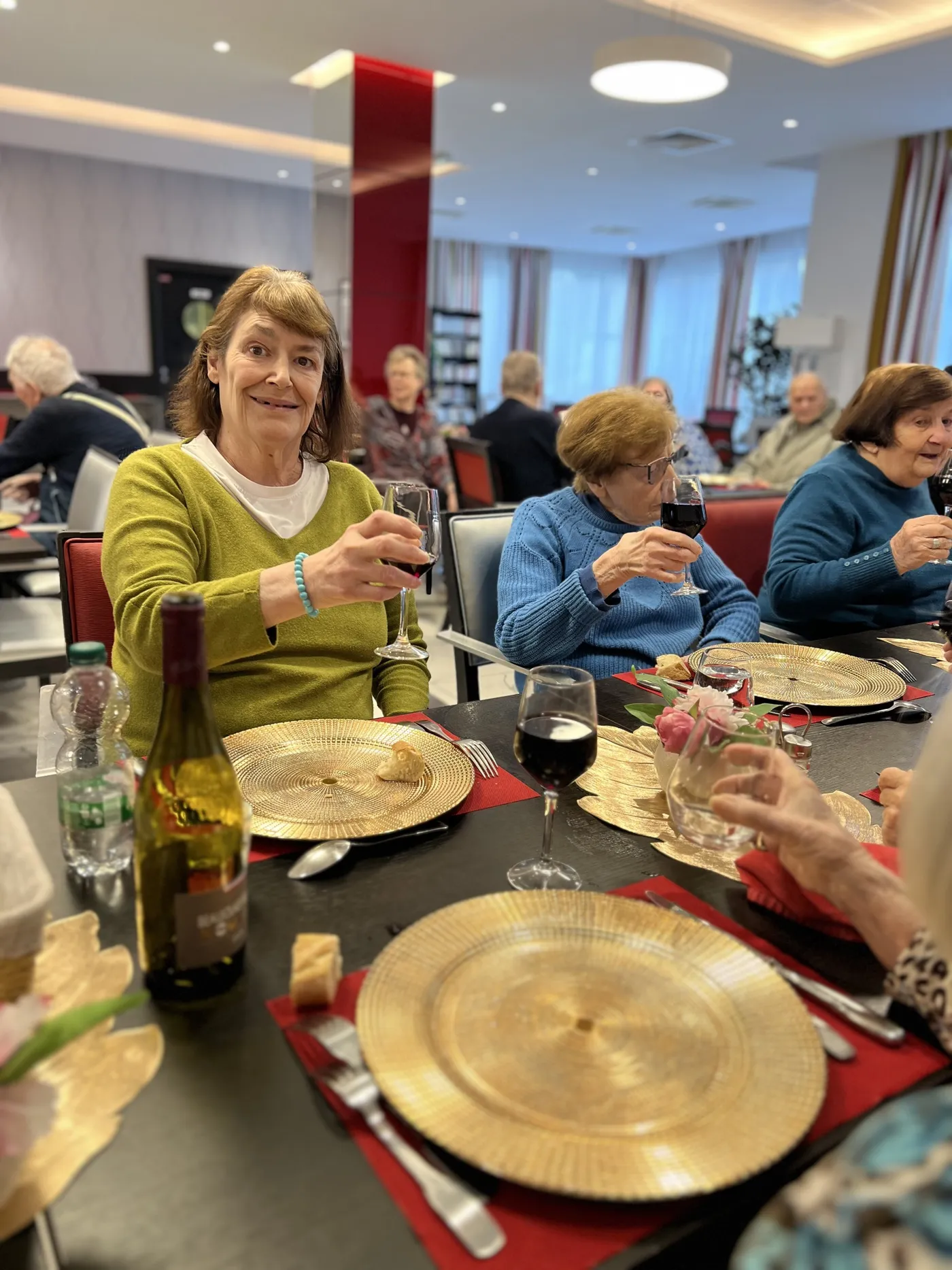 Une résidente en pull vert anis tient son verre de vin à hauteur de poitrine et s'adresse à l'objectif, la table dressée d'assiettes dorées devant elle