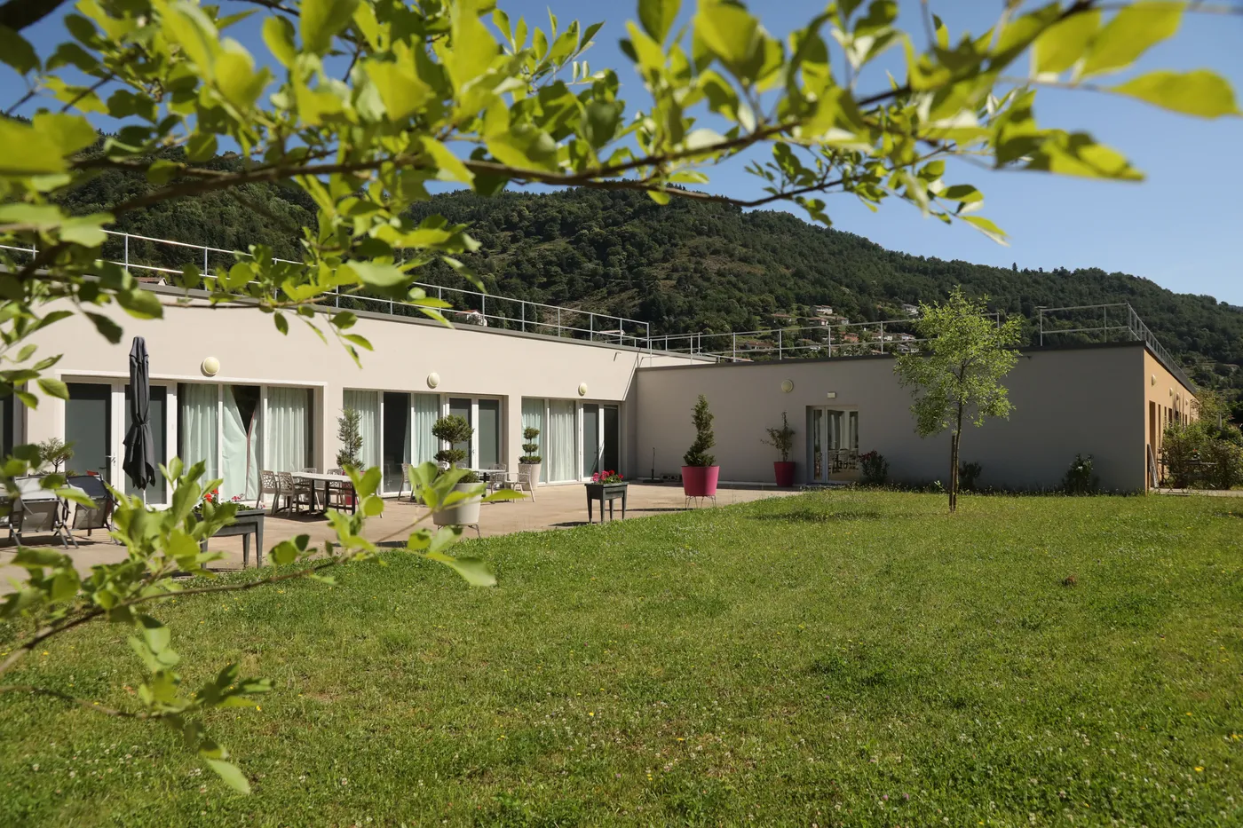 La résidence du Cheylard et sa terrasse vues depuis la pelouse, encadrées par des feuillages
