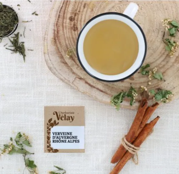 Une tasse de tisane vue de dessus sur une planche en bois, avec des bâtons de cannelle et un sachet de verveine