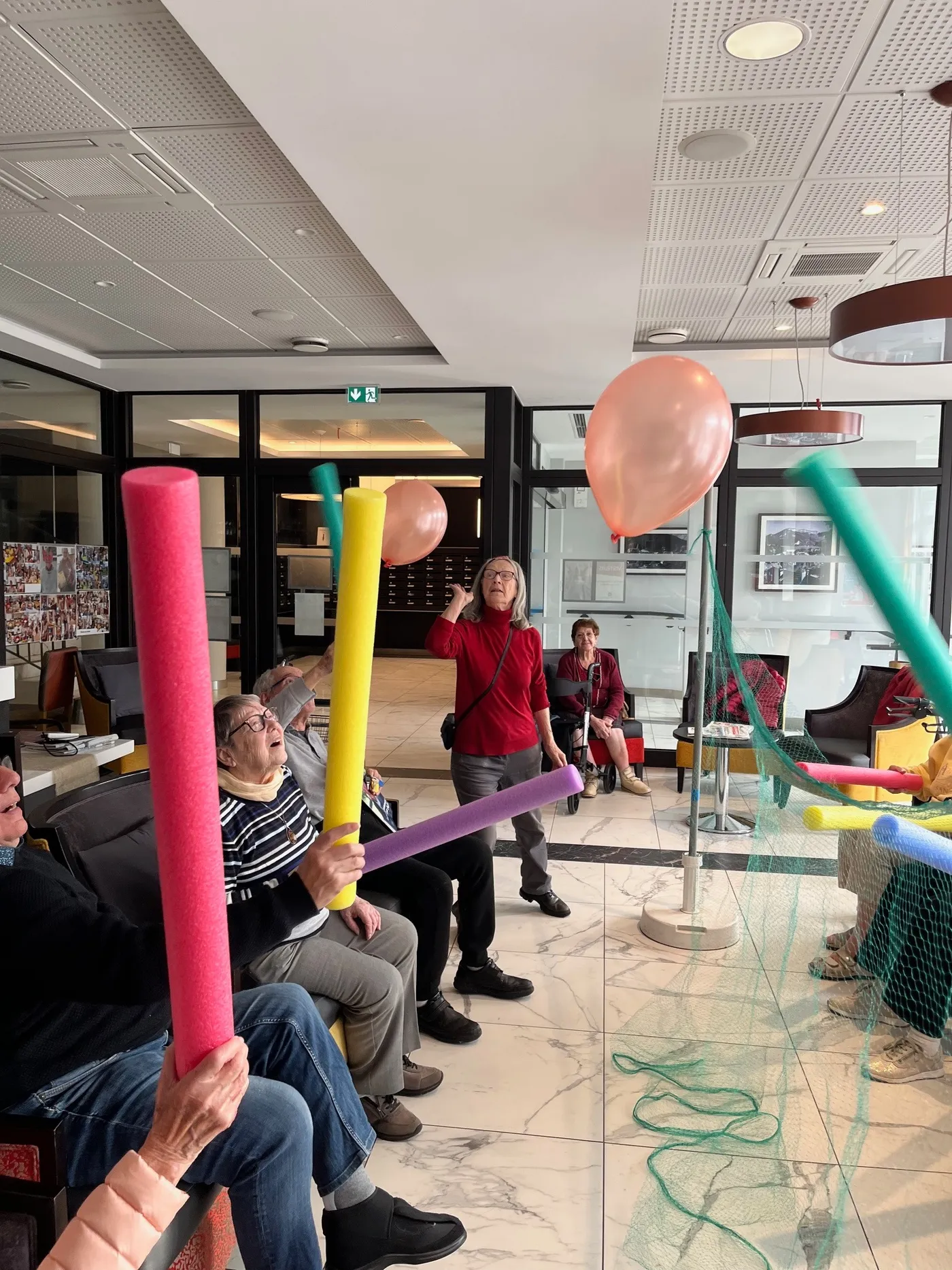 Résidents assis en cercle brandissant des frites en mousse colorées, un ballon en l’air, l’animatrice au centre