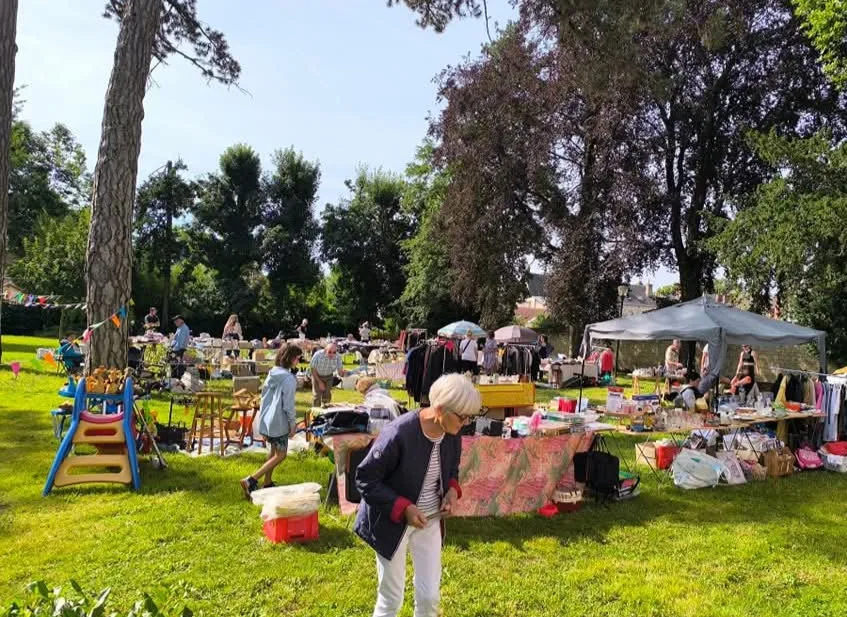 Brocante installée sur la pelouse d’un parc arboré : stands sous barnum, portants de vêtements et tables d’objets anciens, visiteurs qui chinent au soleil.