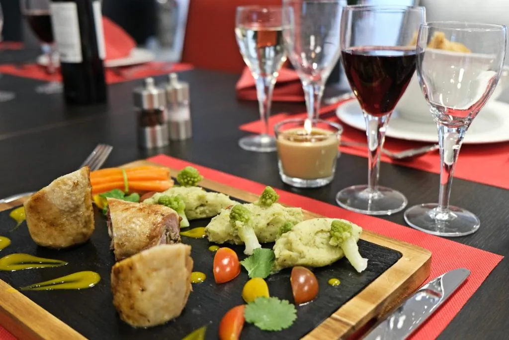 Assiette dressée : viande, carottes, brocolis et tomates cerises sur ardoise, verres de vin en arrière-plan