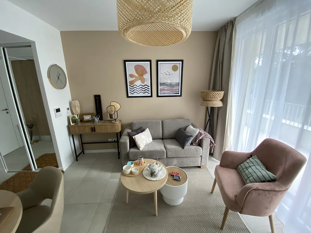Séjour d’un appartement témoin de Toulouse-Guillaumet : canapé gris, fauteuil rose, tables basses rondes et grande baie voilée